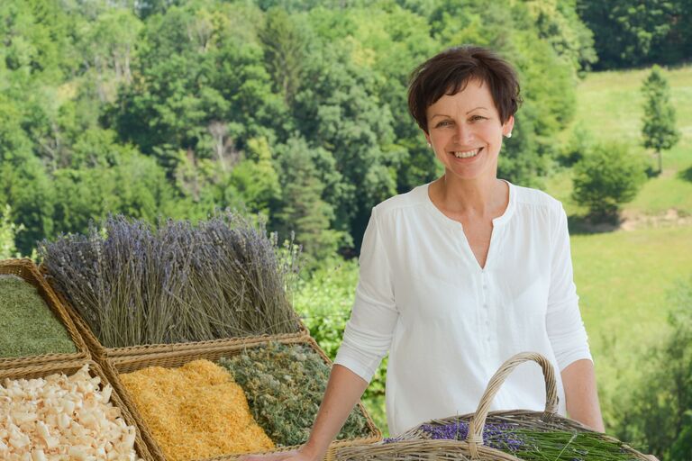 Eine Frau Präsentiert frische Kreuter in Körben, im Hintergrund ein Waldstück mit Wiese